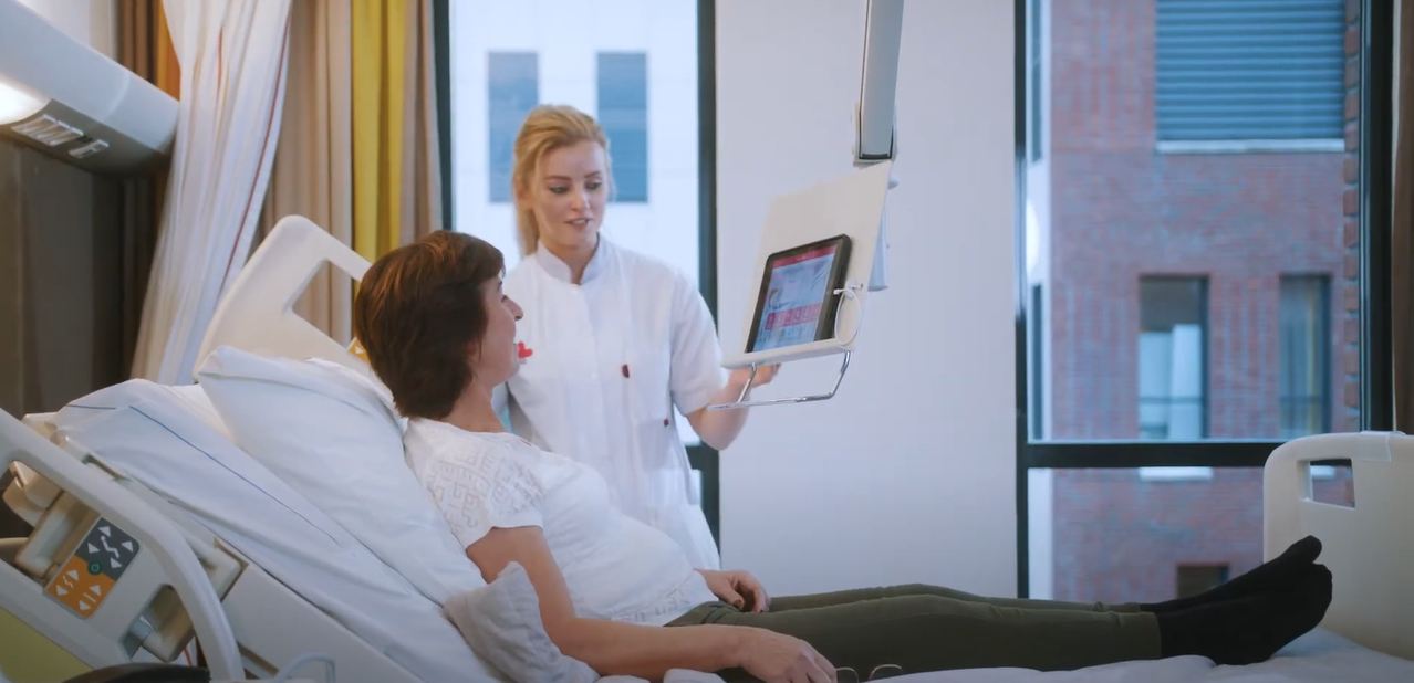 A nurse in a white uniform stands by a hospital bed showing a tablet to a  patient lying in the bed. A professional medical facility setting.